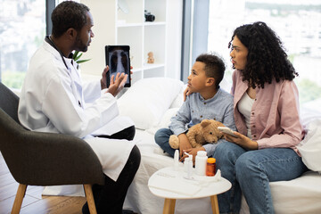 African male doctor showing x-ray to worried African mother and young son in hospital. Child holding teddy bear while sitting on bed surrounded by medicines.