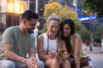 Three friends laughing and using a smartphone outdoors in the city, showcasing the joy of friendship and technology in the summer