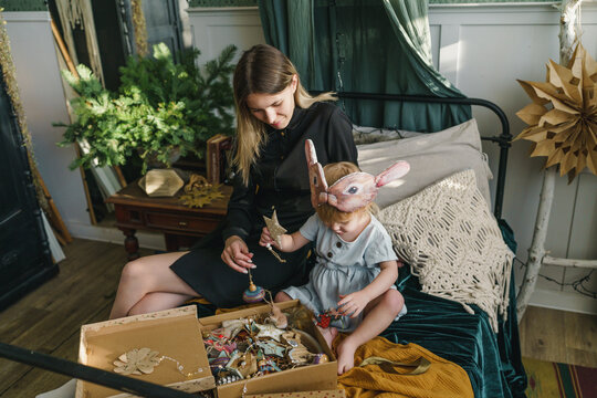 Happy mother and daughter sitting with Christmas ornaments in box on bed at home
