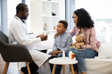 African male doctor examines young boy with concerned African woman comforting him. Doctor offers medicine for recovery at medical check-up.