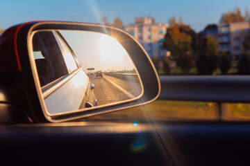 The sun is reflected in the mirror, evening road. View in the side rear view mirror of a red car driving along the highway and street
