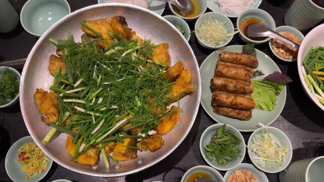 Vietnamese fried spring rolls Cha Gio, with a dish of boneless grilled hemibagrus fish, river catfish fried called Cha Ca La Vong Hanoi, table of diners with fresh dill and spring onion, cilantro