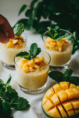 mango sago pudding in glass cups, with a white tablecloth background. Mint leaves and yellow mint are placed on the side. Fresh mangoes are cut into small pieces next to the cups. 