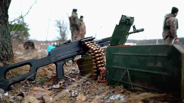 On the ground in a field there is a light machine gun with cartridges against the background of soldiers. Close-up of machine gun cartridges. Small automatic weapons. Light machine gun. Firearms
