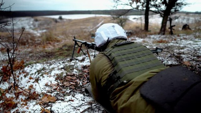 A machine gunner fires a machine gun on a military field. Many spent machine gun cartridges are on the ground. A machine gunner lies on the cold ground with a firearm. Military training