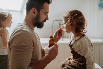 Sick girl with fever and cold at home. Father giving his daughter medicine on spoon, cough syrup or...