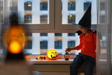Little girl sits by window in witchs hat and halloween pumpkin lantern in autumn evening. Child at...