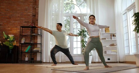 Cheerful Indian Asian young couple joyfully practicing the standing Virabhadrasana yoga pose at home, showcasing strength and balance together