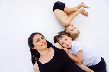 A young pregnant woman with her cute sons in casual clothes are lying on a white floor. An attractive mom is having fun with her boys at home. © Dasha Petrenko