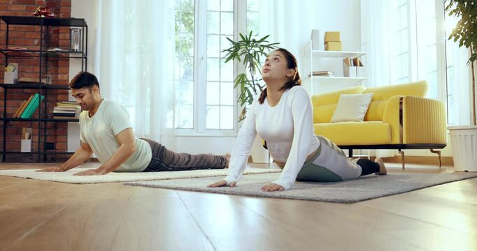 Indian Asian young couple learning yoga in the comfort of their lavish home, practicing bhujangasana or back bend pose together to enhance flexibility and improve spinal strength
