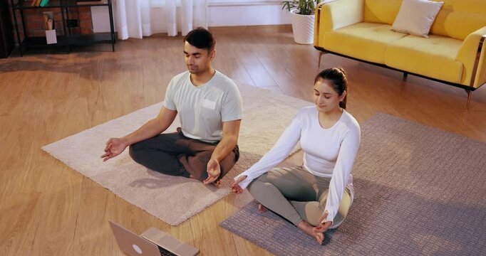 Indian Asian young couple happily following an online yoga class, performing meditation, pranayam, and breathing exercises together while seated in their lavish home