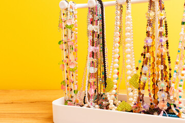 Different handmade chains of beads, pearls and natural stones hanging on a stand in front of yellow background. Collection of fashionable accessories.