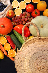 Round straw bag and various seasonal fruits and vegetables on dark background. Summer and fall produce. Top view.