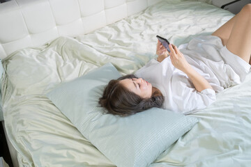 A woman lying comfortably on a bed, smiling while focused on her smartphone and tablet. The bright and cozy atmosphere captures a cheerful moment of happiness in her personal space.