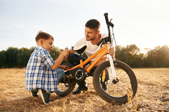 Sitting and repairing the bike. Dad with son are with bicycle on the field outdoors