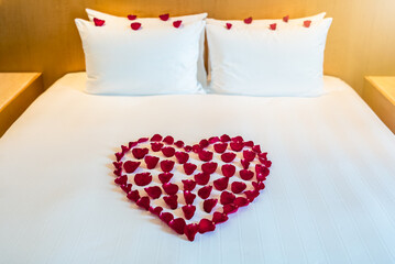 A heart shaped arrangement of red rose petals decorates a bed with white pillows