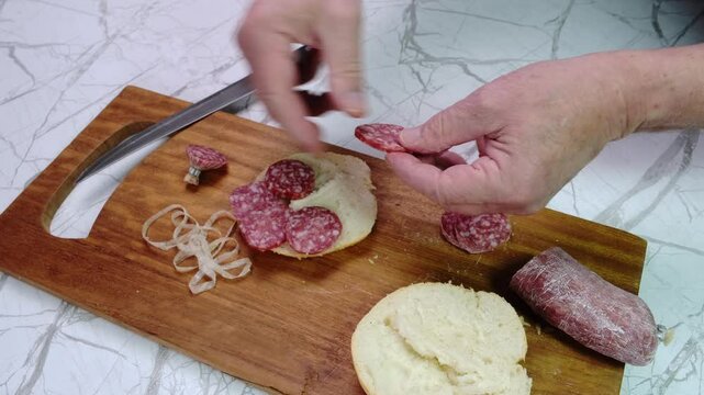 Uomo prepara il panino con le fette di salame, tagliere e coltello sul tavolo