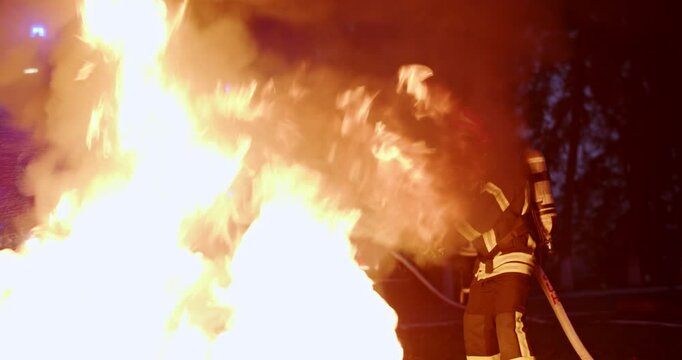 Courageous fireguard in full equipment and helmet standing outdoor at night and fighting with fire. Open flames outside in evening. Brave fireman extinguishing fire with water in dark.