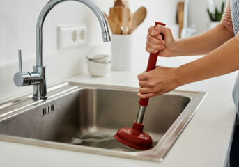 Person unclogs a kitchen sink using a plunger in a modern kitchen with minimalist decor during daylight