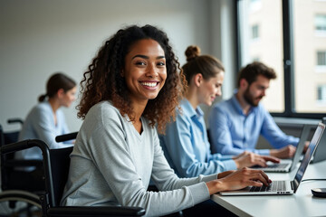 A young African American woman with wavy shoulder-length hair in a light gray sweater sits in a wheelchair and works at a laptop against the background of colleagues