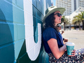 Woman wearing wide brim hat sitting against colourful blue tone wall looking out and holding coffee
