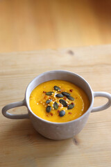 Bowl of creamy pumpkin and potato soup, with pumpkin seeds and chilli flakes toppings. Selective focus, wooden background.
