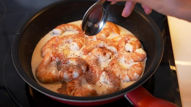 Close-up of woman hand pouring spices with a tablespoon to the raw peeled shrimps stewing in cream in a black pan on electric stove 