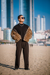 A Korean guy in a leopard fur coat walks along the beach against the backdrop of the autumn landscape of Busan Haeundae.