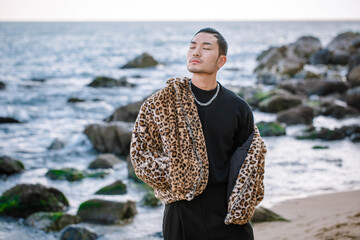A Korean guy in a leopard fur coat walks along the beach against the backdrop of the autumn landscape of Busan Haeundae.