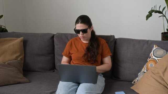 Zoom in view of visually impaired young woman with black glasses using laptop on couch in living room