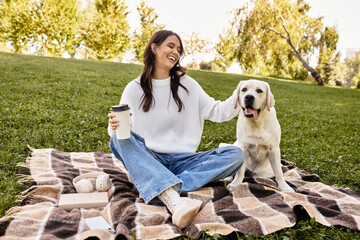 A young woman smiles while seated on a blanket, savoring her coffee alongside a happy dog.
