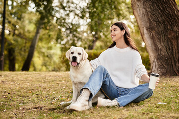 A young woman in cozy autumn attire relaxes on the grass with her cheerful dog, sipping coffee.