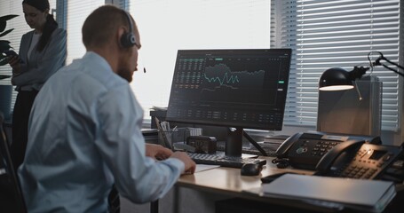 In the stylish modern office: Male financial trader in headset works on computer, talks to business client on call, monitors stocks and share market, takes notes. International stock exchange company.