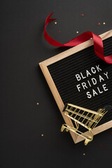 Black Friday sale signboard with a miniature shopping cart and red ribbon on dark vertical background, representing holiday shopping and discounts © ActionGP