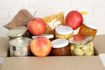 Different food products for donation in box near white brick wall, closeup
