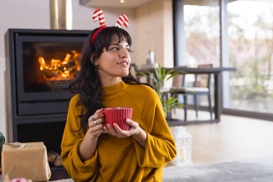 Christmas time, woman enjoying hot drink by fireplace, wearing festive headband at home