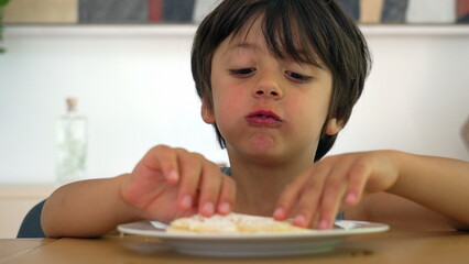 Young boy eating tapioca at the dining table, taking a large bite while holding the food with both hands, enjoying a relaxed meal in a home setting