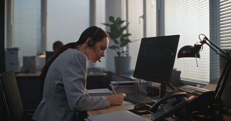 In stylish modern office: Female financial trader in headset works on computer, talks to business client on call, monitors stocks and share market, takes notes. International stock exchange company.
