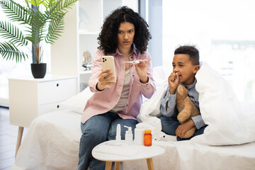 African mother checks temperature of sick boy lying on bed during online doctor consultation by phone. Boy holds teddy for comfort, wrapped in blanket. Scene reflects family care and remote healthcare