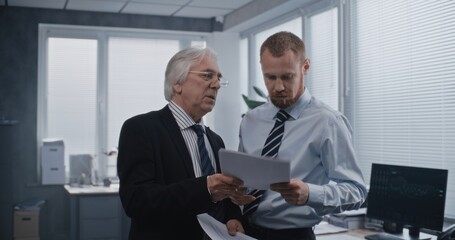 In modern office of global financial company: Senior director and young manager or financial analyst talking, discussing business strategy of company, analyzing papers and documents. Businesspeople.