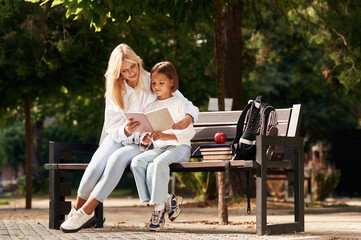 Reading together. Little schoolgirl with her mother are sitting on the bench outdoors