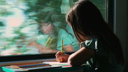 Young girl drawing with a pencil on a moving train, her reflection visible on the window as she focuses on her creative activity with the countryside blurred