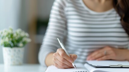 A person is seated at a table, writing in a notebook, with a decorative plant nearby, dressed in a striped top.
