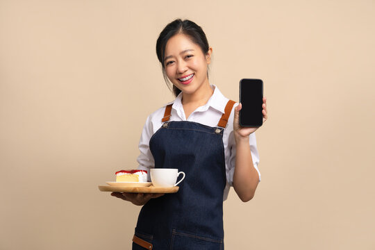 Asian female waitress in apron holding bread and coffee drink on wooden tray with smartphone on beige background.