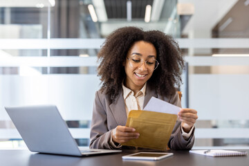 A smiling young African-American businesswoman is sitting at the desk in the office and opens an...