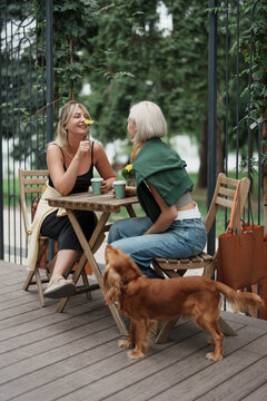 Two women are sitting at a cafe table outside, laughing and holding coffee cups, with a cute dog nearby. The atmosphere is relaxed and friendly.