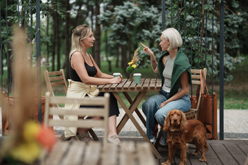Two women enjoy a conversation at an outdoor cafe. A brown dog sits beside them, creating a relaxed, friendly atmosphere. They are surrounded by nature in a tranquil setting.