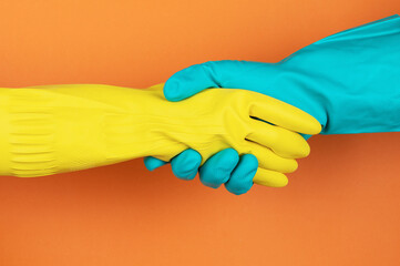 Two hands wearing yellow and blue rubber gloves shake hands against a bright orange background,...