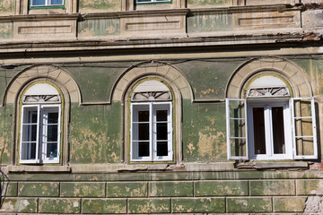 A weathered building facade in Sibiu, Romania, featuring three arched windows with white frames and shutters. The building is painted in a muted green and has peeling paint in several areas. 