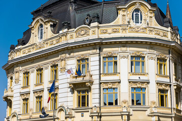 A close-up of a historic building facade in Sibiu, Romnania, featuring a curved window surrounded by intricate decorative elements. The building has a dark roof and a light-colored exterior.